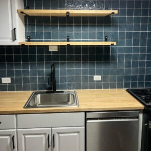 Kitchen with dark blue tile backsplash, butcher block countertop, floating wood shelves and a black faucet